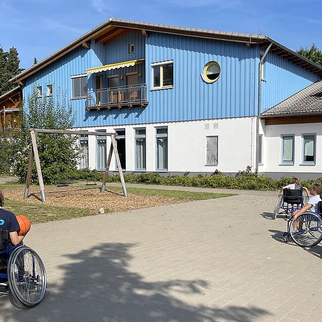 Vier Kinder im Rollstuhl spielen Rolli Basketball vor dem Drehpunkt Gebäude