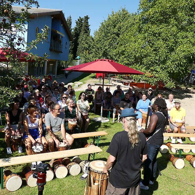 Die Besuchende im Drehpunktgarten mit Trommeln zwischen den Beinen hören konzentriert den Anweisungen zu.