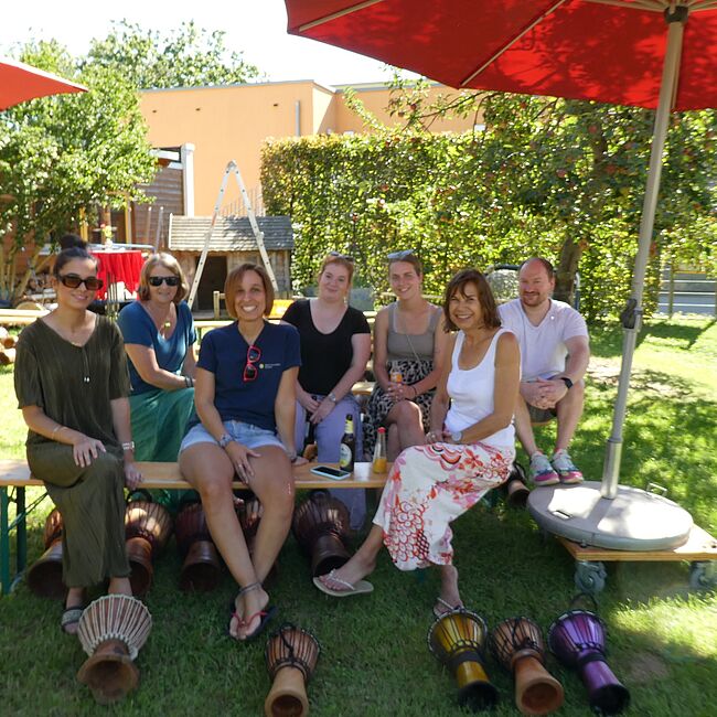 Ein Teil des Drehpunktteams sitzt im Drehpunktgarten unter einem Schirm und machen ein Gruppenbild mit Trommeln.