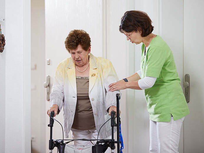 Ambulanter Pflegedienst Ambulanter Pflegedienst: Dame hilft älterer Dame mit Rollator beim Betreten einer Wohnung.
